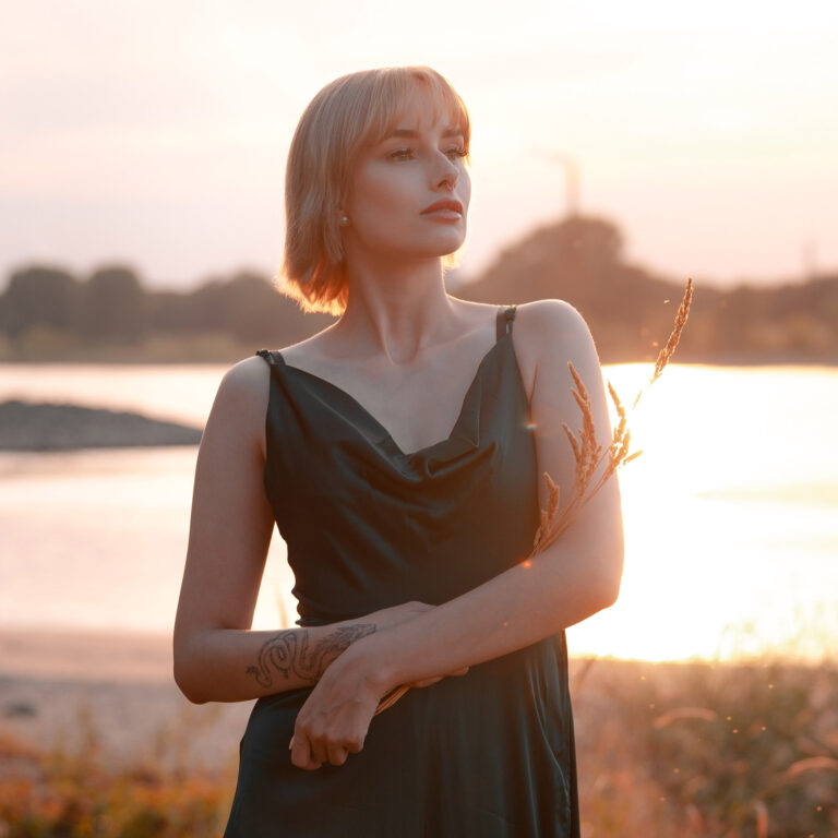 Stimmungsvolles Porträt einer Frau im eleganten Kleid bei Sonnenuntergang am Fluss – Du strahlst Selbstbewusstsein und Ruhe aus, während das warme Licht deine natürliche Schönheit in der Naturkulisse hervorhebt.